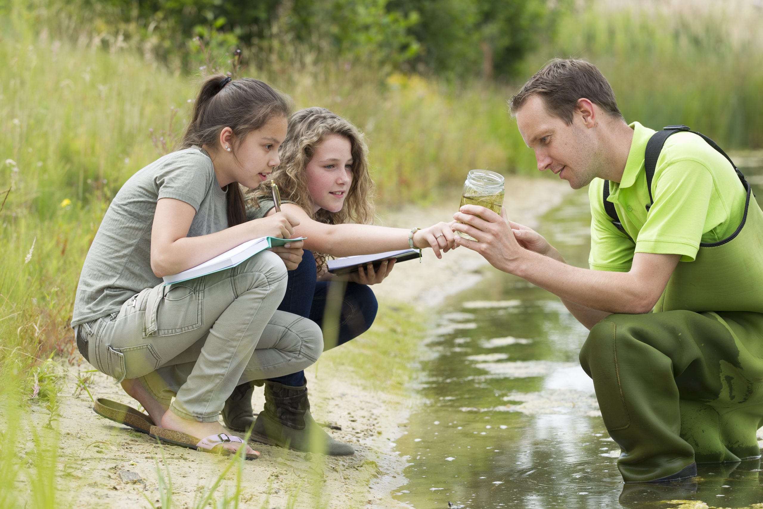 A man in waders squats in the edge of a body of water, showing something to two girls who are crouched by the edge of the water.